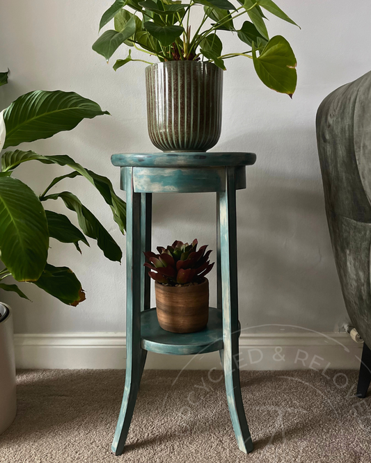 A distressed teal blue green wooden end table with a single shelf, styled with a potted green plant on top and a small succulent in a pot on the shelf, set against a neutral indoor background. n upcycled piece by Surrey upcycler Jodi Walker