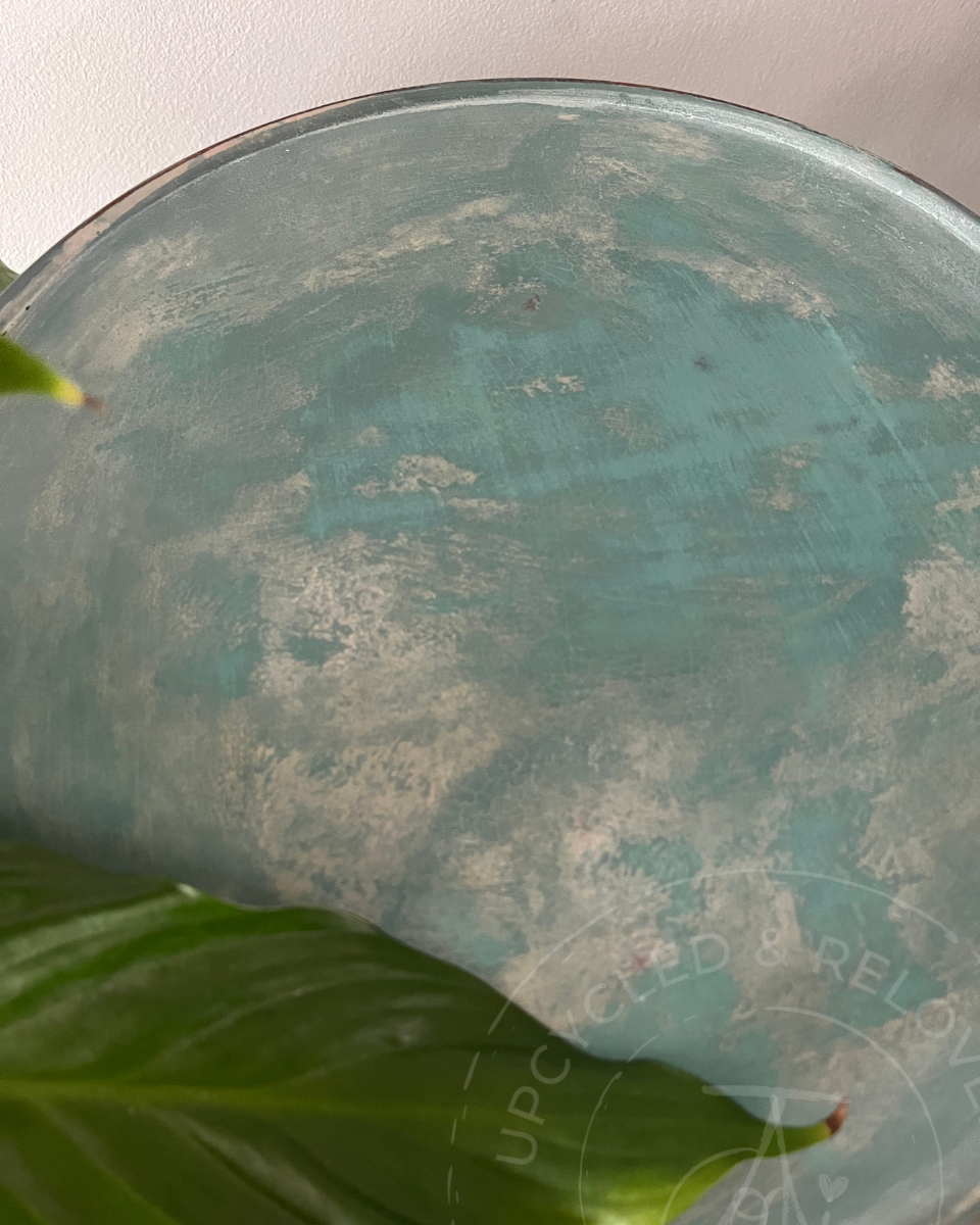 A close-up of a round, distressed teal plant stand table top with a mottled, weathered finish, painted in Fusion Milk Paint and upcycled to a smooth buttery finish with a natural aged look