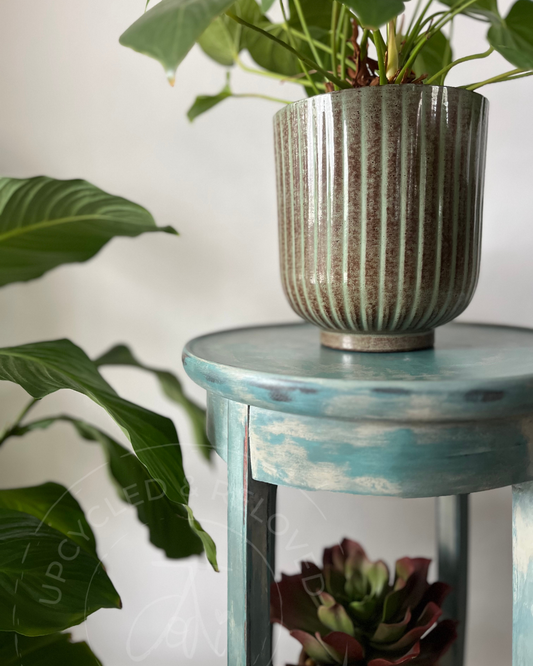 A plant pot on a wooden upcycled side table plant stand finished in blues, greens and beige with a distressed, aged look and a soft sheen. A unique refinished shabby chic piece of furniture by Jodi Walker