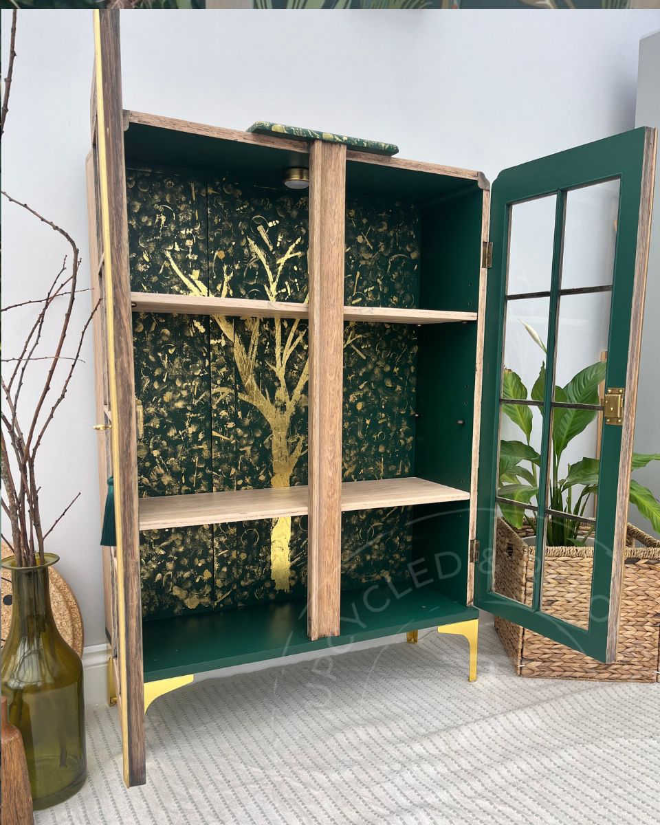 Side on view of a tall, light wooden cabinet with a green painted glass panelled door and gold legs, shown open with shelves and a decorative forest-patterned interior panel. The interior is a rich green with a gold leaf tree and gold patterns