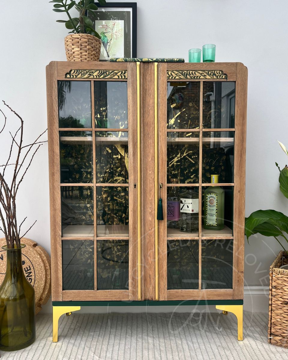 A tall wooden double-door drinks cabinet in a pale oak with glass panes, gold-toned decorative trim, and gold legs, styled in a home interior with plants, bottles, and decor. A unique upcycled piece of furniture, the item is one of a kind