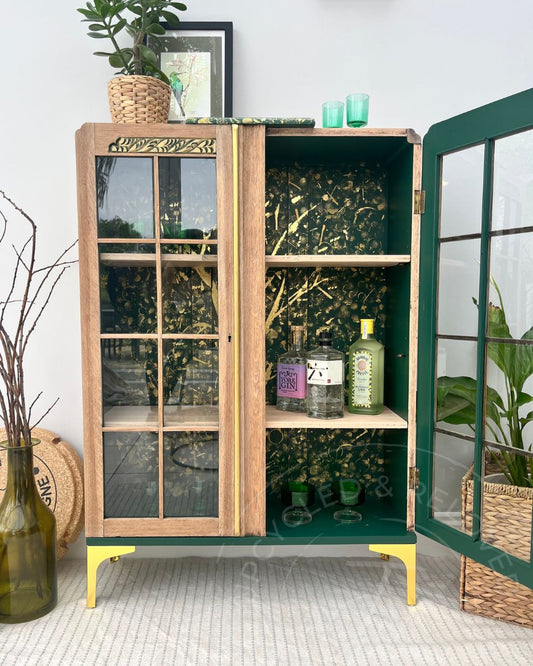 A solid oak wood display or drinks cabinet with a green painted frame and interior, featuring glass-paneled doors and open shelves. The cocktail cabinet interior is painted forest green with hand painted gold tree and forest style pattern. The oak cabinet has modern gold legs