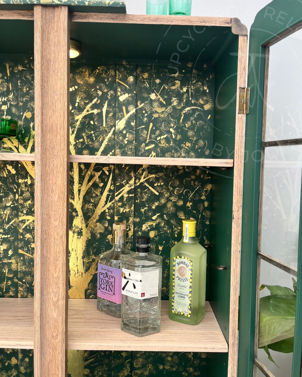 A upcycled wooden shelving cabinet with a dark green interior featuring a gold tree pattern, holding several bottles including a gin bottle and a green liqueur bottle. An unique upcycled piece by Jodi