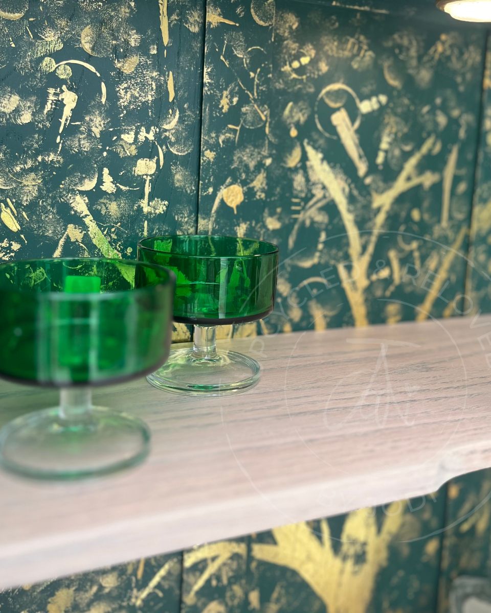 The interior of an upcycled drinks cupboard showing two green glass goblets on a light oak wood shelf in front of a dark green and gold hand painted abstract patterned backing. The watermark bears the logo "Upcycled & Reloved by Jodi" - A furniture artist based in Surrey