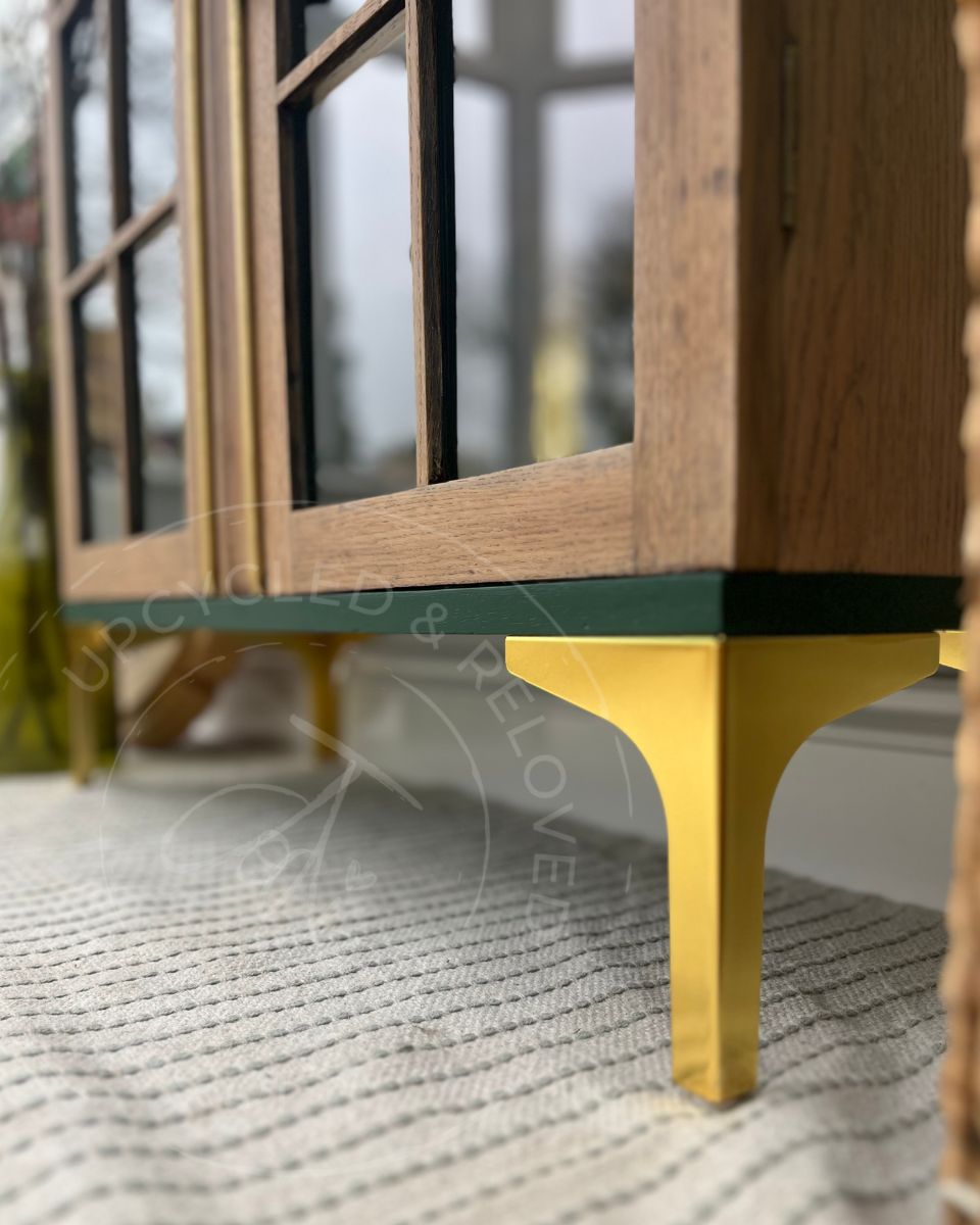 Close-up of an oak wood drinks cabinet with modern gold legs ,glass panels, a dark green base trim shown on a textured light rug indoors.