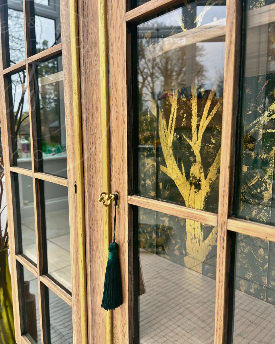 Close-up of a light wood upcycled cabinet with glass panes on the doors, a gold lock and handle, and a dark green tassel hanging from the latch. The interior is visible through the glass showing dark greens with gold  trees painted in the back. The item is an upcycled unique piece
