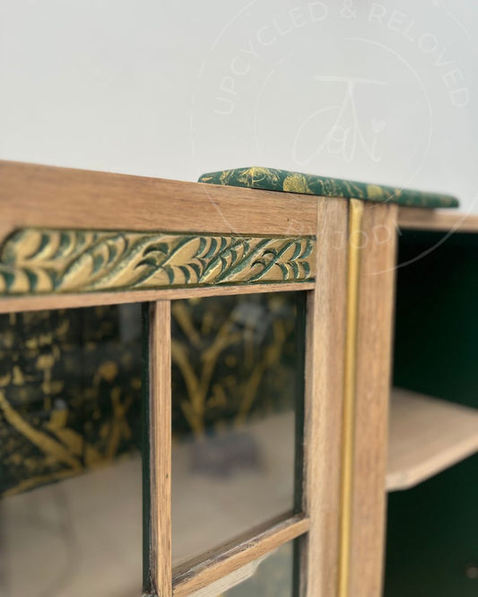 Close-up of a wooden cocktail cabinet door with a deep green and gold pattern on the decorative inlay and top edge. The image shows glass paned doors and a gold door trim. The logo upcycled by Jodi appears as an overlay
