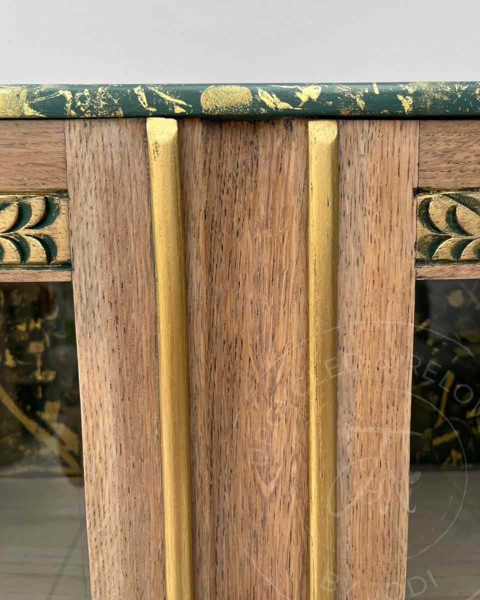 Close-up of an upcycled light oak wooden cabinet top and door trim accented in gold with wood grain panels, gold-toned metal accents, and a dark green top with gold leaf detailing.