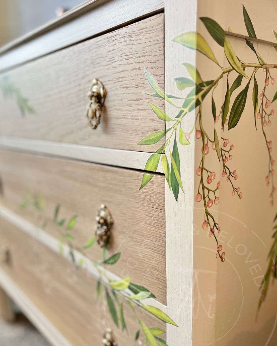 Close-up  side view of a light wood 3 drawer vintage dresser with ornate brass drawer pulls and green leaf and pink berry botanical motifs on a cashmere cream painted background. The logo 'Upcycled & Reloved by Jodi' is visible.