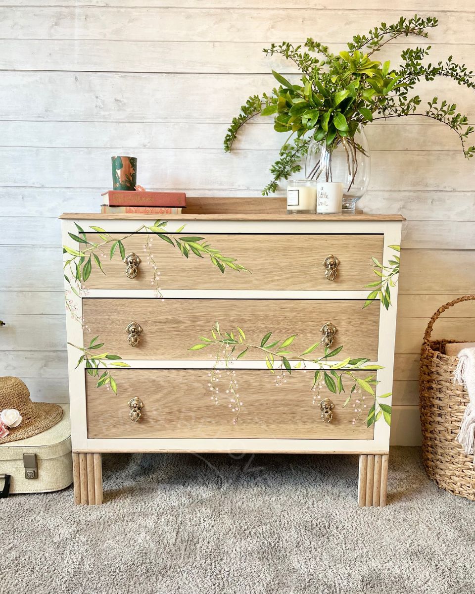 A three-drawer vintage upcycled wooden chest of drawers with a light natural oak wood finish, painted cream drawer fronts, and green leafy vine decals. It is styled in a rustic room setting with a vase of greenery, candles, and a woven basket on a gray rug against a white wood-paneled wall. A unique upcycled piece of furniture