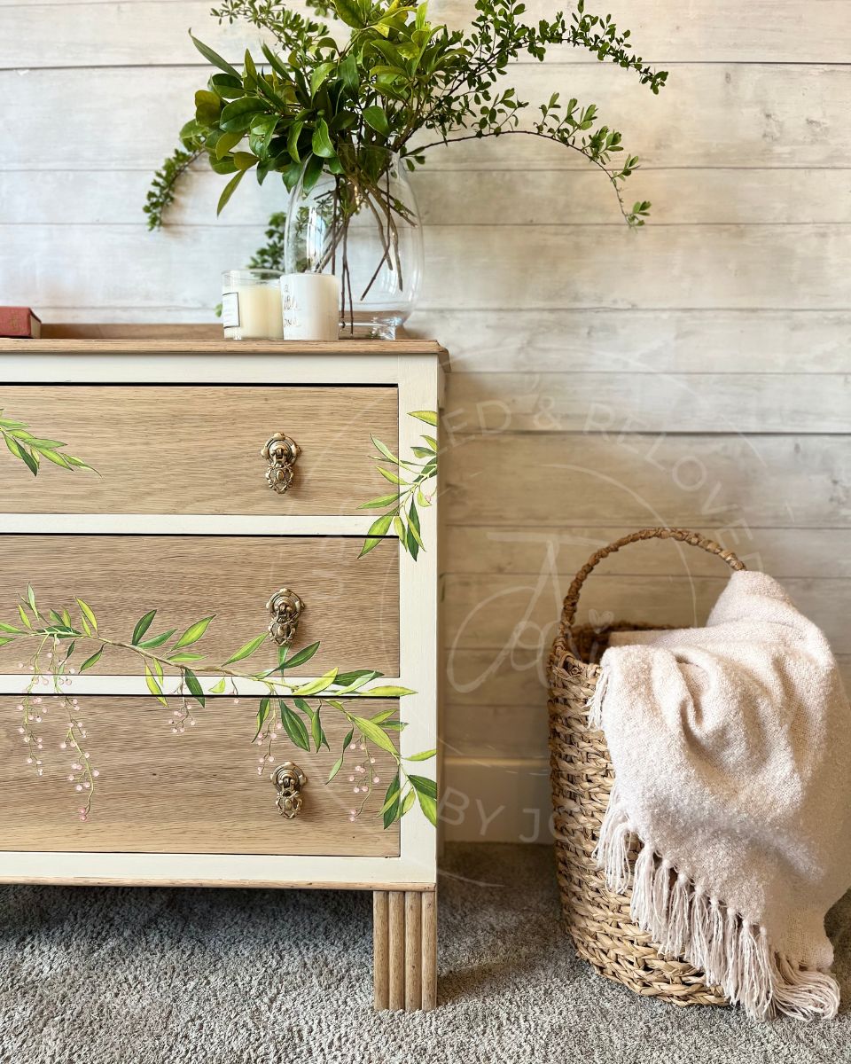 A vintage oak three-drawer wooden chest of drawers with a light  paint washed oak finish and cashmere cream trim, styled in a rustic room next to a woven basket with a cream throw blanket. The drawers have trailing leaves and pink blossoms or berries detailed across the fronts.