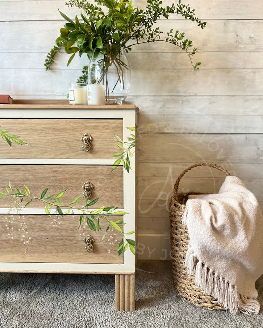 A vintage oak three-drawer wooden chest of drawers with a light  paint washed oak finish and cashmere cream trim, styled in a rustic room next to a woven basket with a cream throw blanket. The drawers have trailing leaves and pink blossoms or berries detailed across the fronts.