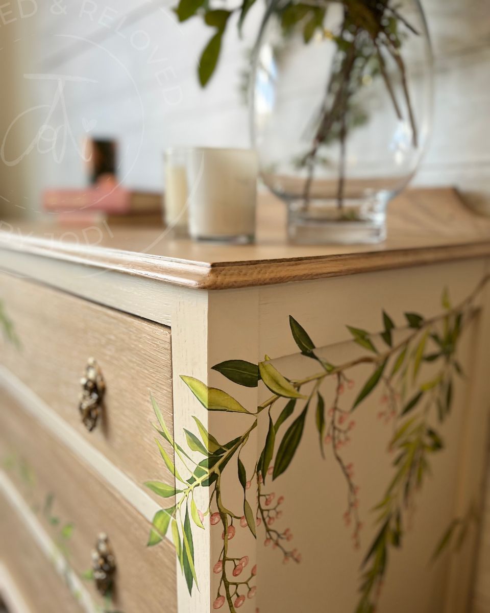 A close-up of a vintage oak wooden dresser with botanical leaf and blossom decals, painted in a soft cream colour, topped with a glass vase and candles in a softly lit interior setting.