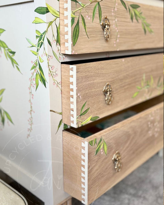 Close-up of a light natural oak three-drawer dresser with green leaf and blossom transfer and brass drawer pulls, set against a creamy white background. The drawers show the original dovetails painted in the cream paint colour