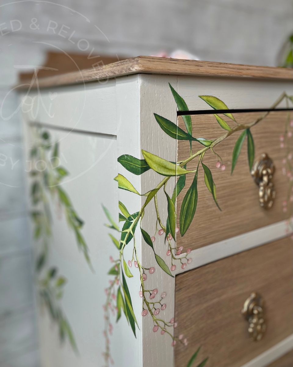 A close-up of a vintage oak chest of drawers with light oak wood grain drawers, decorated with green leafy vines and small pink flowers, featuring ornate original vintage brass handles. A unique refinished piece of furniture