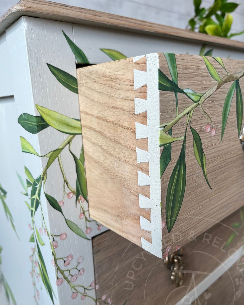 An open drawer of a vintage oak chest with natural light oak wood grain drawer fronts, cashmere whiter painted base and leaf blossom botanical transfer. The logo says Upcycled & Reloved by Jodi' a Surrey based furniture upcycler