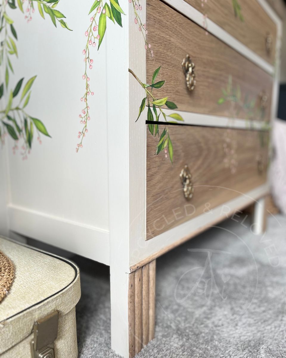 A close-up  side on view of an upcycled oak wood chest of drawers with vintage handles and legs, a neutral creamy white painted side panel and delicate green floral vine decals, featuring brass drawer pulls and standing next to a vintage suitcase