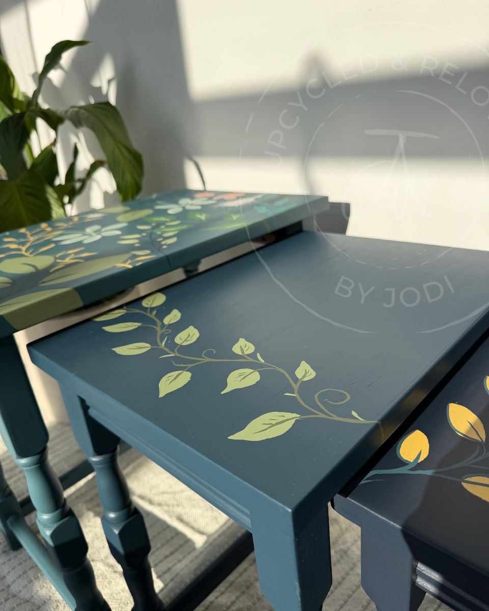 A set of three nested tables painted in shades of deep teal blue, to a lighter mid-blue decorated with hand-painted light green leafy vines and yellow floral accents. The upcycled tables are arranged in an indoor setting with natural sunlight casting shadows on a white wall and a green plant in the background.