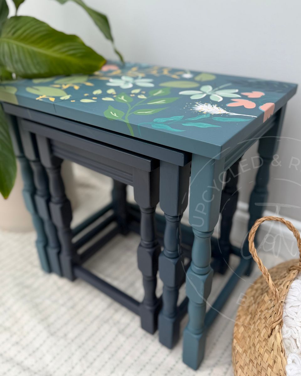 A set of  painted wooden stacking tables with a seaside blue top featuring a hand-painted botanical floral pattern, and solid carved legs. The nesting tables are painted in 3 shades of blue from medium blue to mid-blue and dark navy blue on the smaller tables.The tables are styled in a home setting with a potted plant and a woven basket.