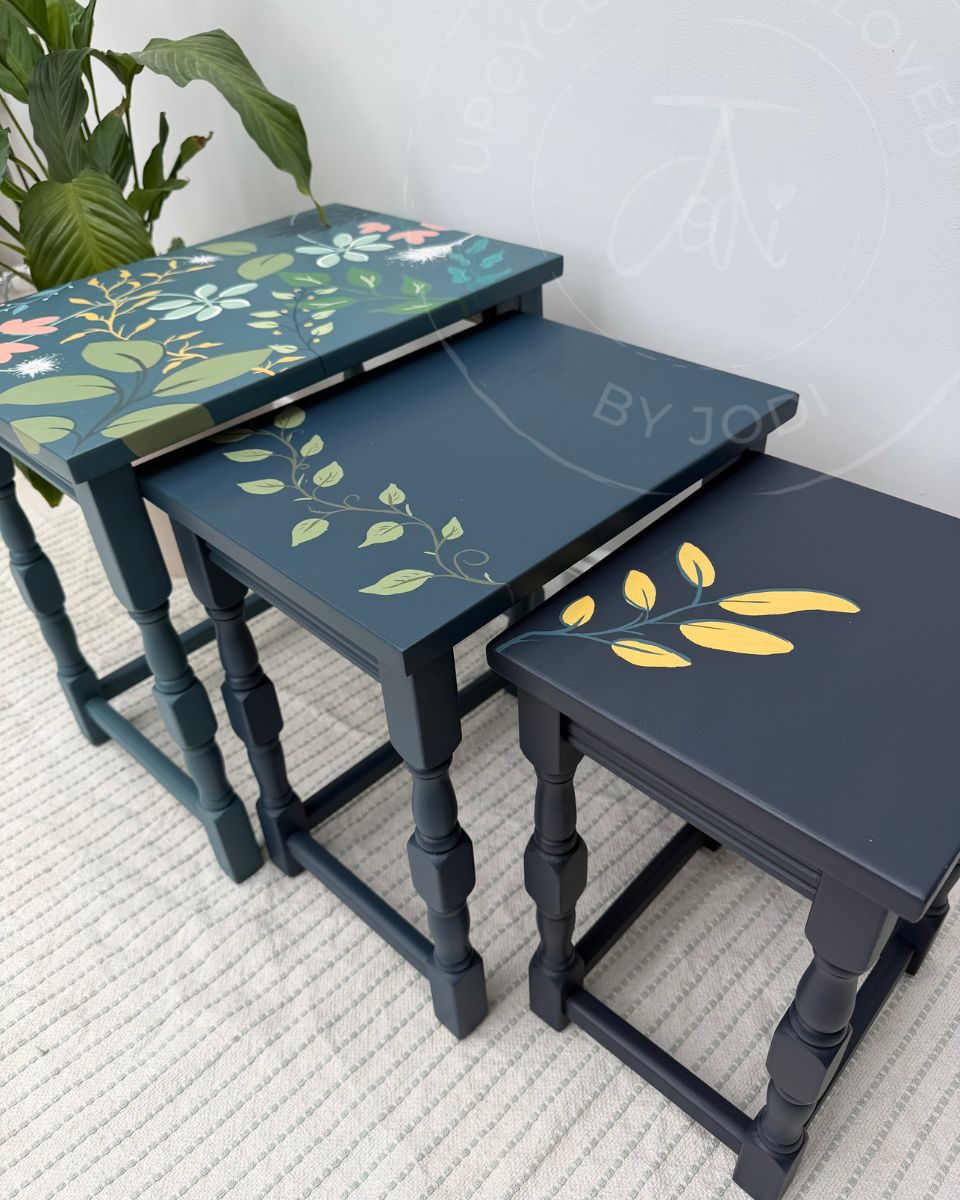 Three nested rectangle tables with turned carved legs, painted in 3 blue teal-blue colours , decorated with hand-painted floral and leaf patterns in green, yellow, pink, and white. The tables are arranged on a textured light beige rug with a green plant in the background. 
