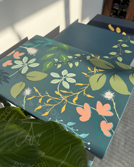 A close-up of the top of a side table painted in Seaside blue Fusion Paint, featuring a hand painted botanical pattern with green leaves, yellow vines, peach flowers, and white dandelion-like blooms on a dark teal background. Two smaller side tables are visible , one with a vine leaf design. The side tables are in blue shades, and a faint script watermark is visible across the surface with thew words UPCYCLED & RELOVED BY JODI