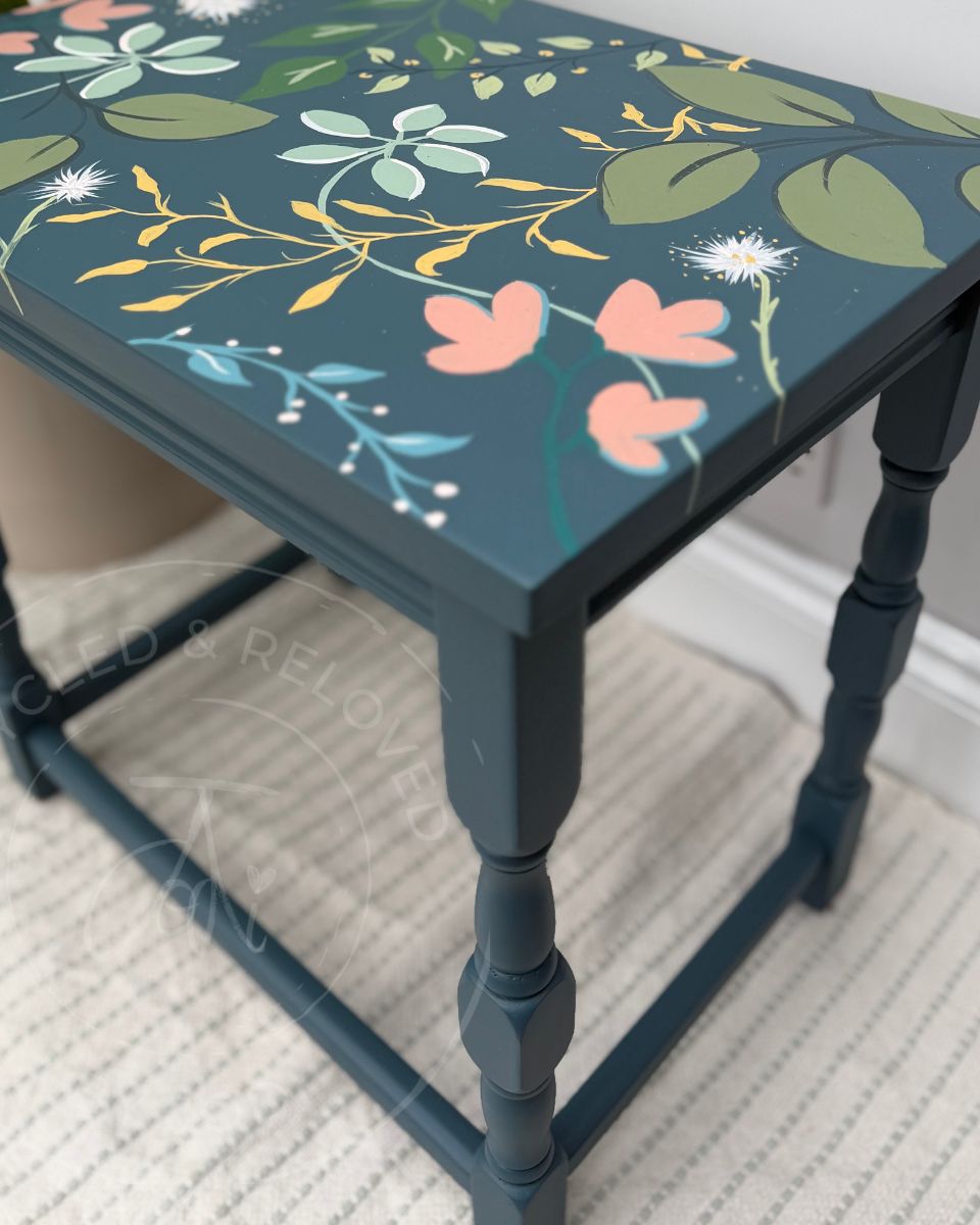 A hand painted wooden side table with a dark teal-blue base painted in Fusion Mineral Seaside Blue with a hand-painted floral pattern featuring green leaves, pink flowers, yellow botanicals and light blue accents, shown in a home setting on a textured rug.