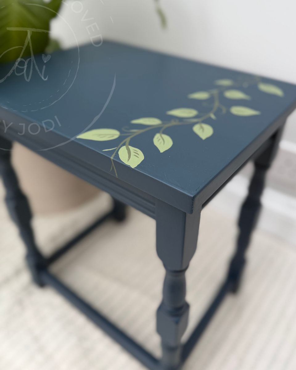 A medium sized side table part of a nest of tables painted in Midnight Blue with a painted green leaf vine design on the top surface, shown in a home setting.