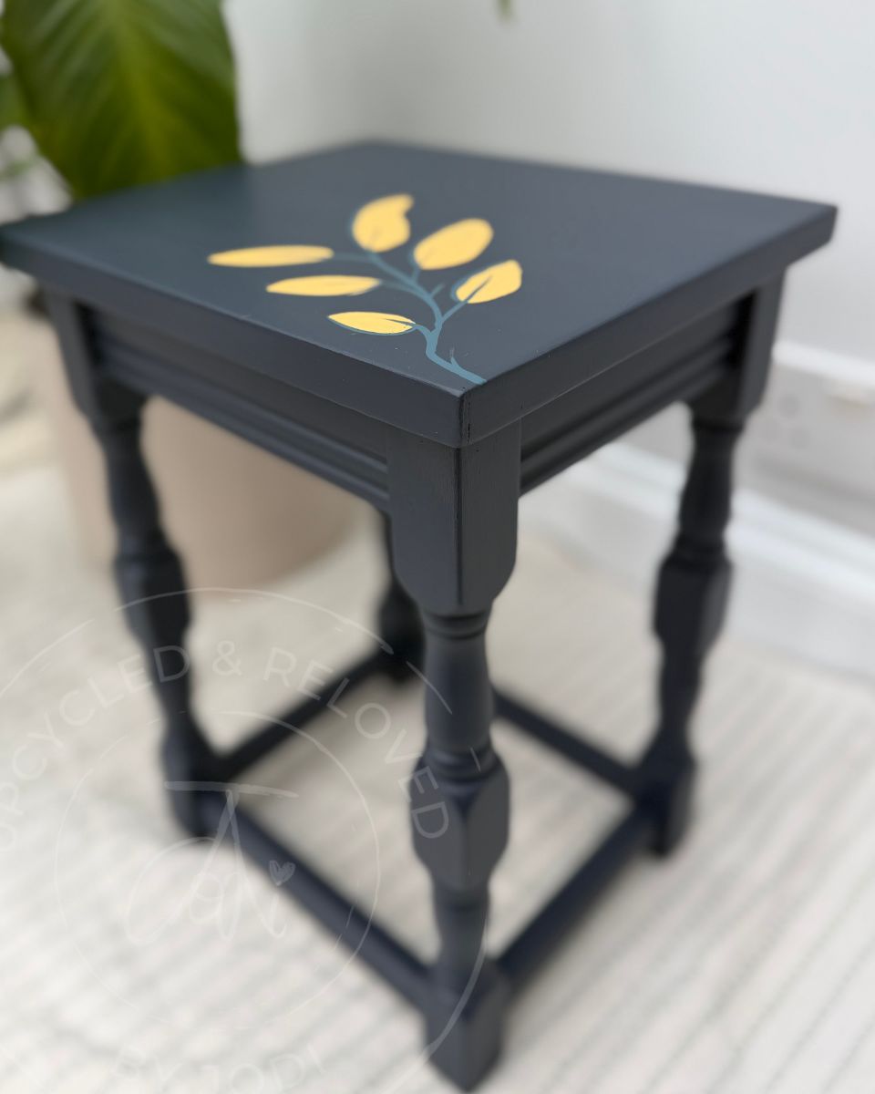 A small navy blue wooden side table with turned legs and a painted yellow and blue leaf motif on the top, part of a set of 3 nesting tables, shown in a home setting with a blurred plant in the background.
