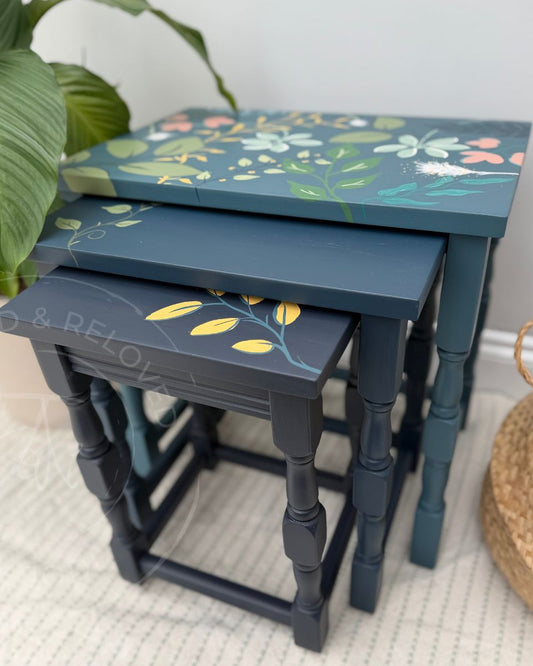 Three upcycled nested nesting tables with a dark teal-blue painted finish in 3 tones of blue and hand-painted floral and leaf patterns on the tops, styled in a home setting with a green plant and woven basket.
