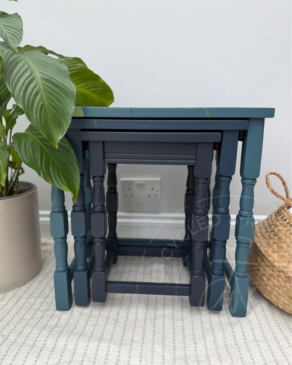 A set of three stacked wooden nesting tables in a rich blue and navy painted finish, styled in a home setting with a potted plant and a woven basket.